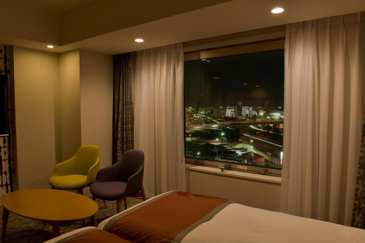 Narita Tobu Hotel Airport Exterior photo The photo shows a hotel room at night. There are two chairs—one yellow and one purple—next to a round table. The room features a large window with curtains that partially cover it, offering a view of a cityscape illuminated by lights. The bed is made